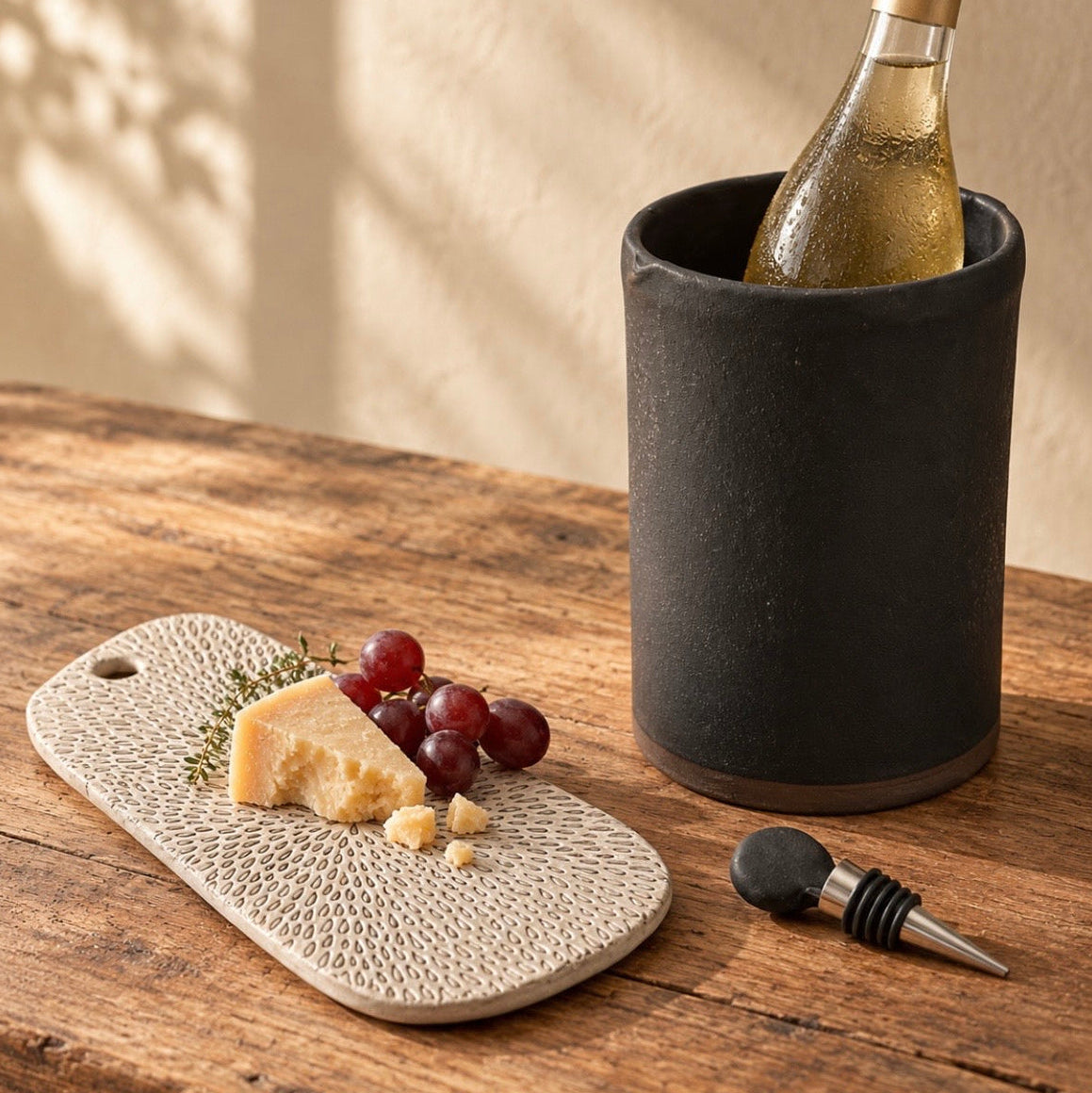 Small Petal ceramic serving board with cheese and red grapes beside a black ceramic wine chiller holding a bottle of white wine on a wooden table.