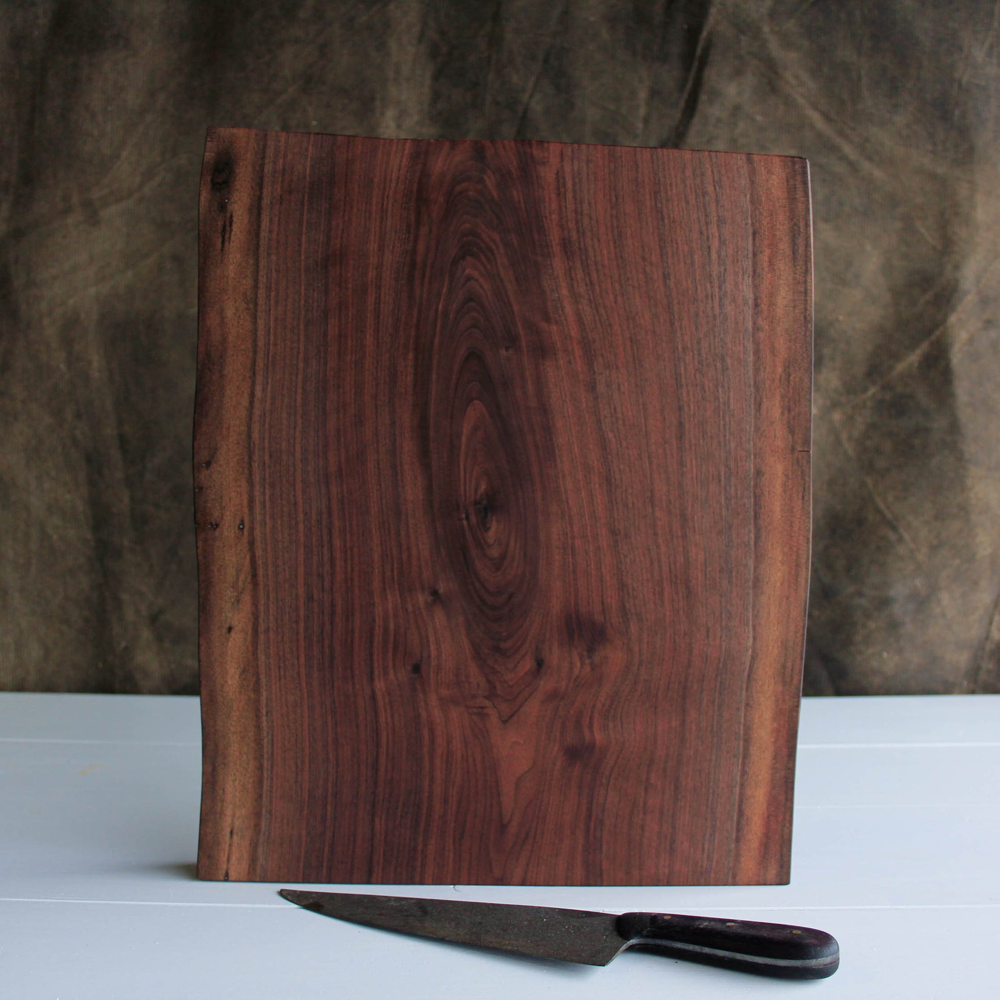 A Picture of a figured walnut cutting board with two live edges standing in front of a kitchen knife