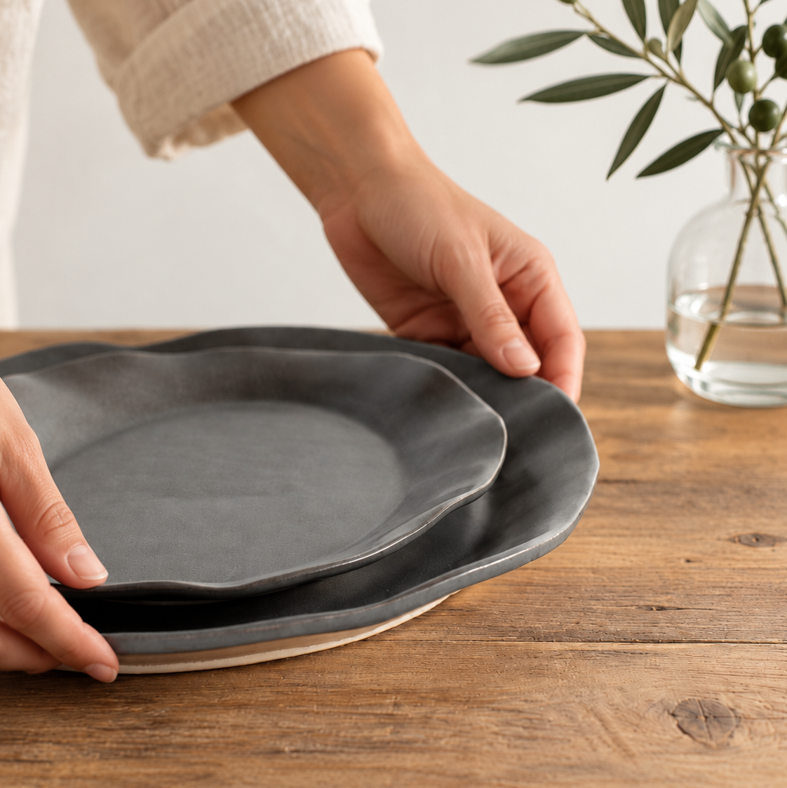 Hands arranging two nested Charcoal Shino oval platters on a wooden table beside a glass vase of leafy branches.
