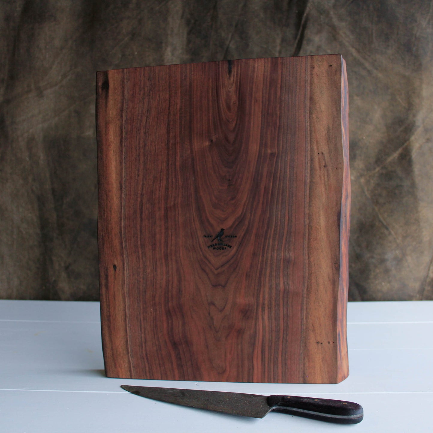 A Picture of a figured walnut cutting board with two live edges and a logo of the meadowlark bird standing in front of a kitchen knife
