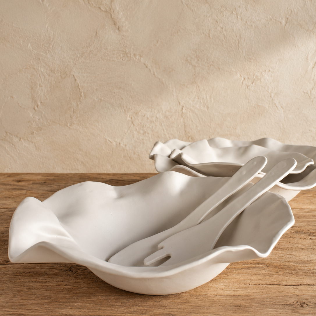 White Matte sculpted serving bowl with ceramic salad servers resting inside, with another rippled-rim bowl behind it on a wooden table