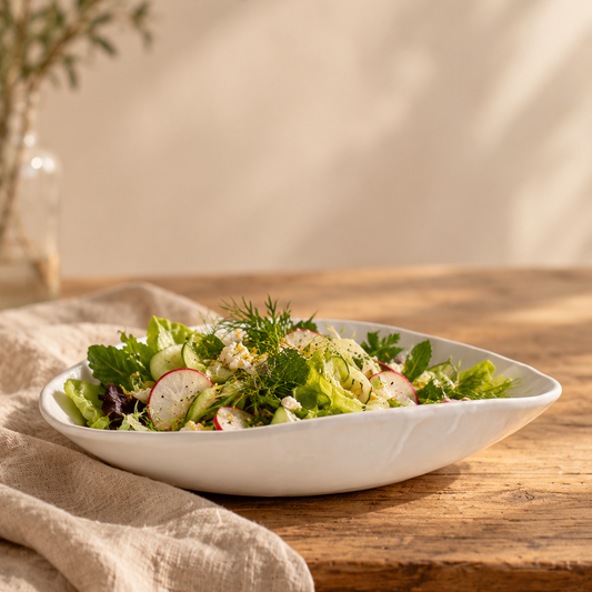 New salad photo: White Matte serving bowl filled with leafy salad, cucumber, and radishes on a sunlit wooden table beside beige linen.
