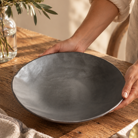 Hands placing a Charcoal Shino serving bowl on a sunlit wooden table, showing its wide rim and shallow profile.