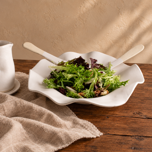 White Matte sculpted bowl filled with leafy salad and ceramic servers beside natural linen on a wooden table.