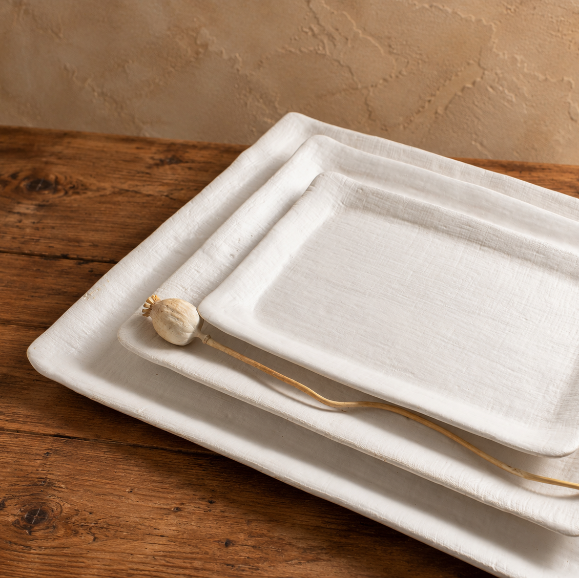 Three nested White Matte linen-textured rectangular stoneware platters with a dried flower stem.
