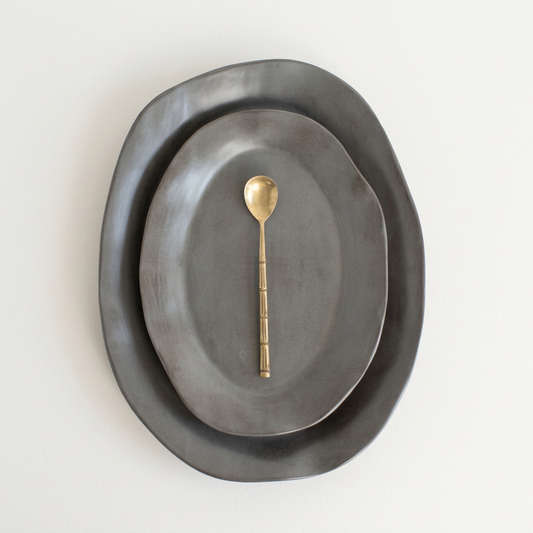Two nested Charcoal Shino oval serving platters with a gold spoon on a porcelain-white background.