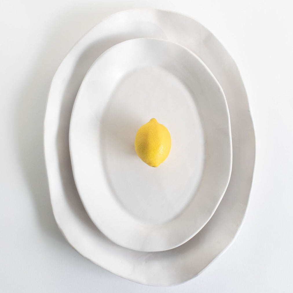 Two nested White Matte oval serving platters with a yellow lemon centered on the smaller platter.