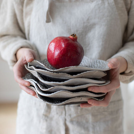 Person in a linen apron holding stacked Petal Square appetizer plates topped with a red pomegranate.